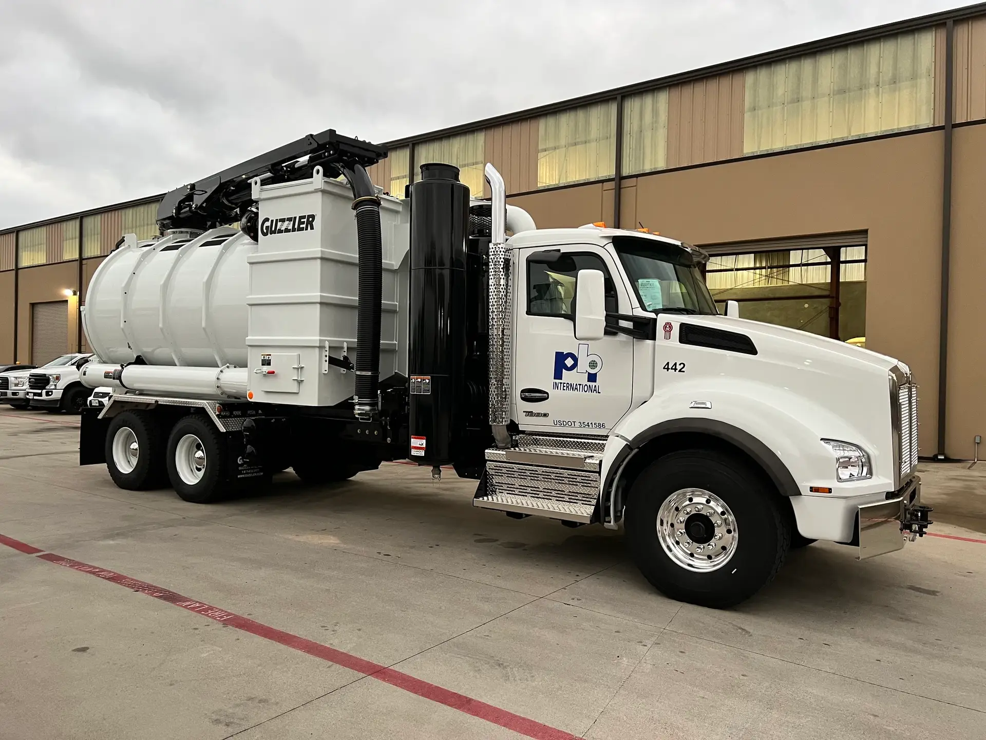 White industrial vacuum truck labeled “Guzzler” with PBI International branding parked outside a warehouse facility.