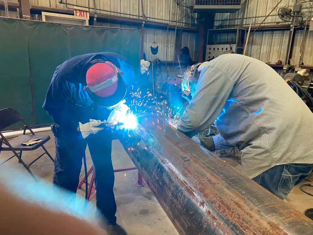 Two industrial workers welding a large metal pipe inside a fabrication workshop with bright sparks flying.