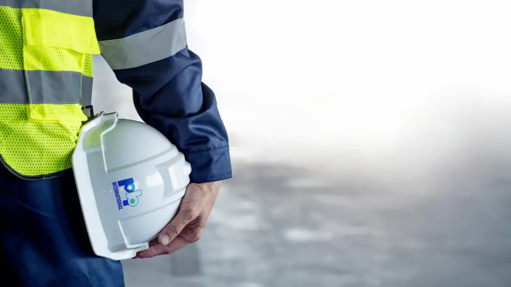 Industrial worker wearing safety gear holding a white hard hat with the PBI International logo.