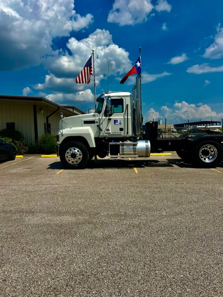 PBI International semi-truck displaying American and Texas flags parked at an industrial facility under a bright blue sky.