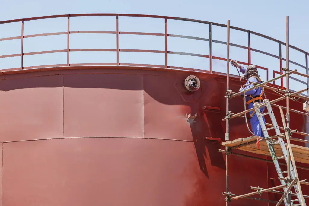 Industrial worker wearing protective gear sandblasting and coating a large metal storage tank on scaffolding.