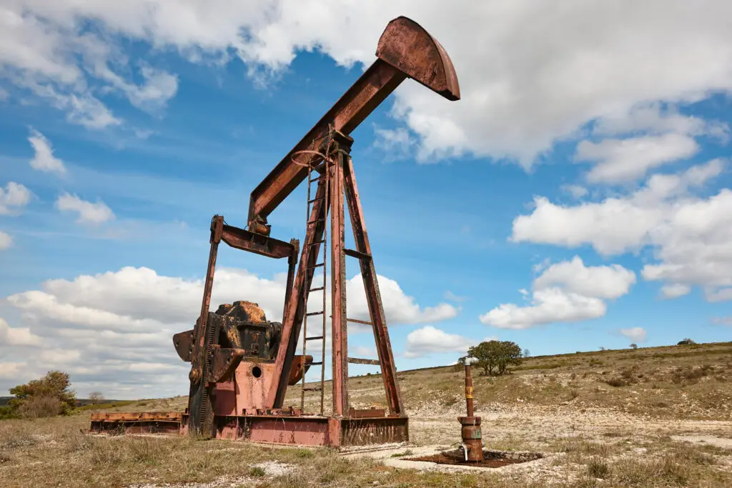Old oil pump jack standing idle in a rural field under a partly cloudy sky.