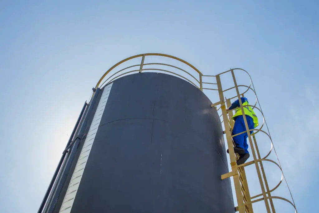 Industrial worker in safety gear climbing a ladder on a large storage tank under clear blue skies.